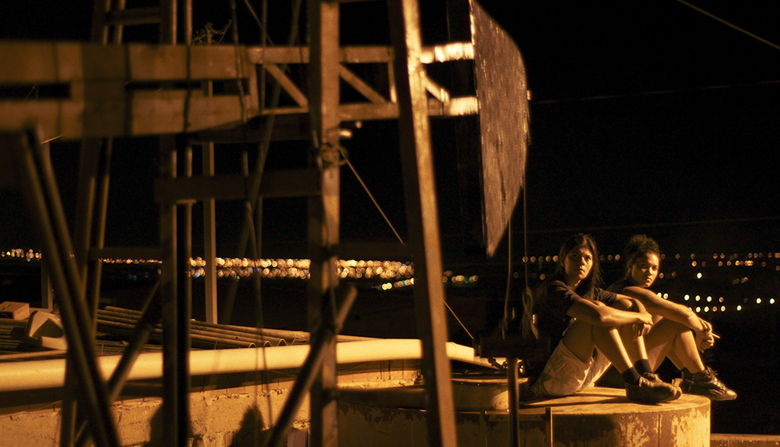 Two women sit near an oil derrick outside, looking up and smoking, at night