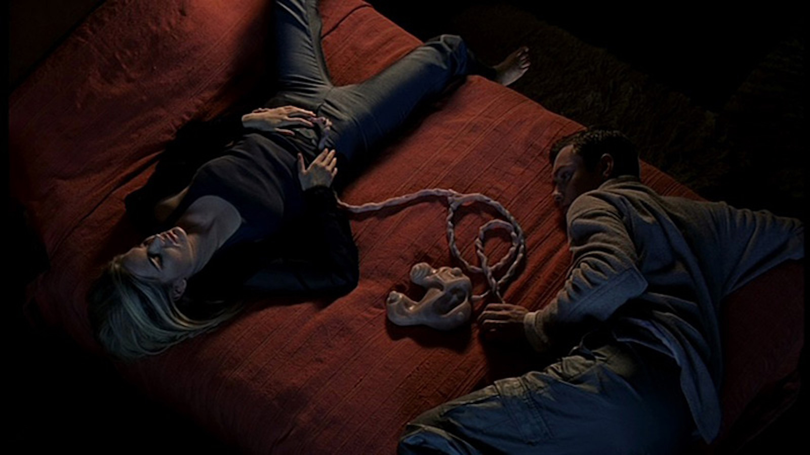 A man and woman lie on a bed apart from each other, connected by fleshy umbilical-like cords in their stomachs