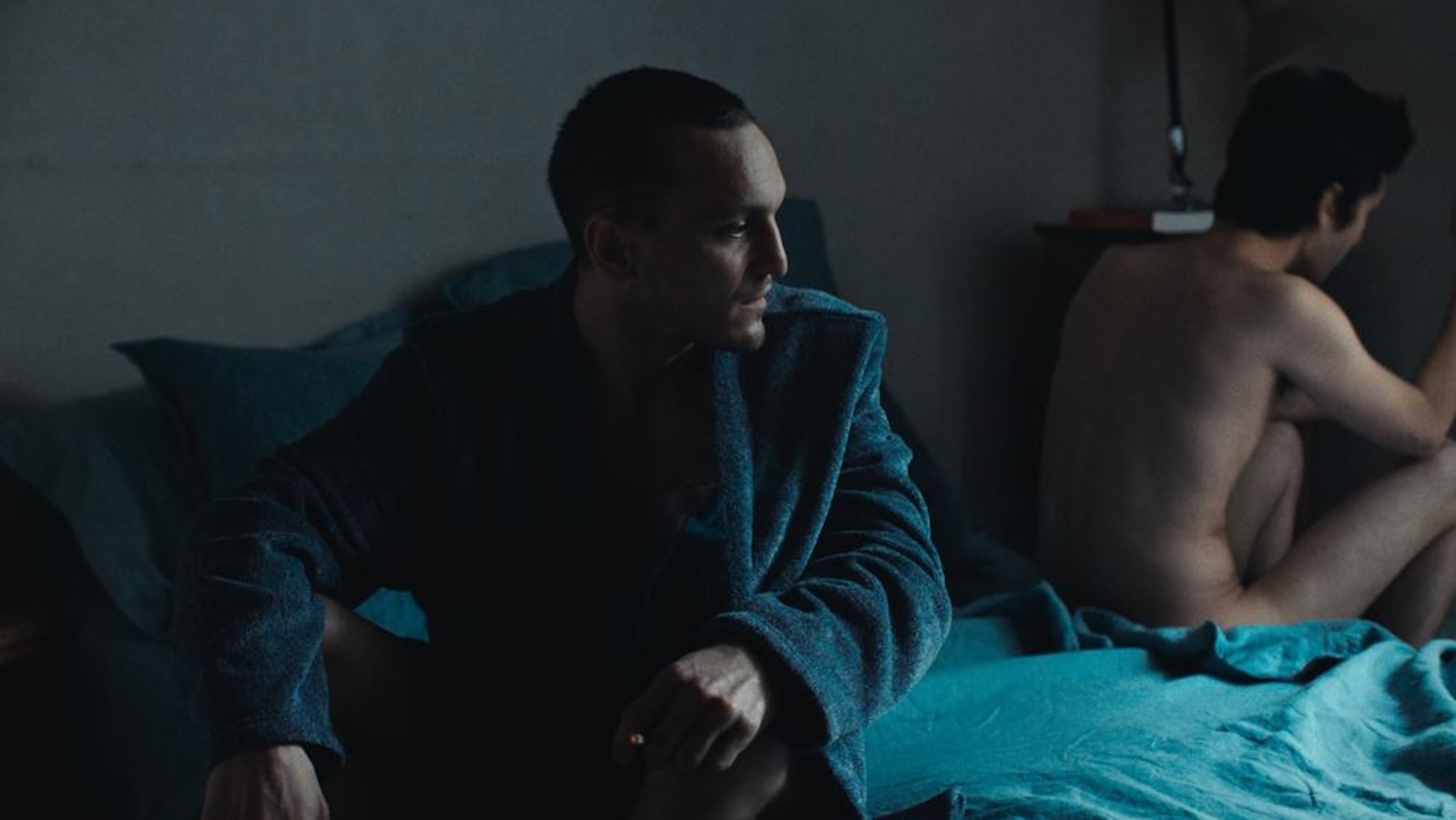 A man wearing a coat sits on a bed turning his head towards a naked man on the other side of the bed, turned away