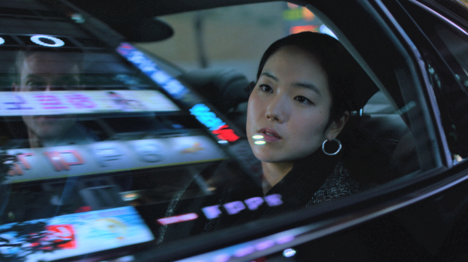 A woman looks out the window from the back seat of a car, with neon city lights reflected in the window
