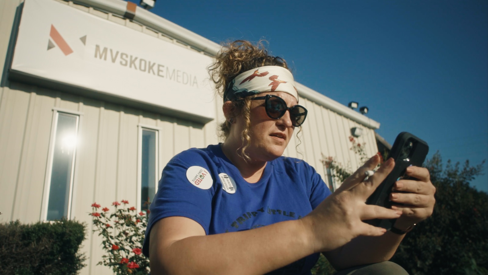 A woman in bandana and sunglasses sits on a curb outside a building looking at her phone and smoking a cigarette