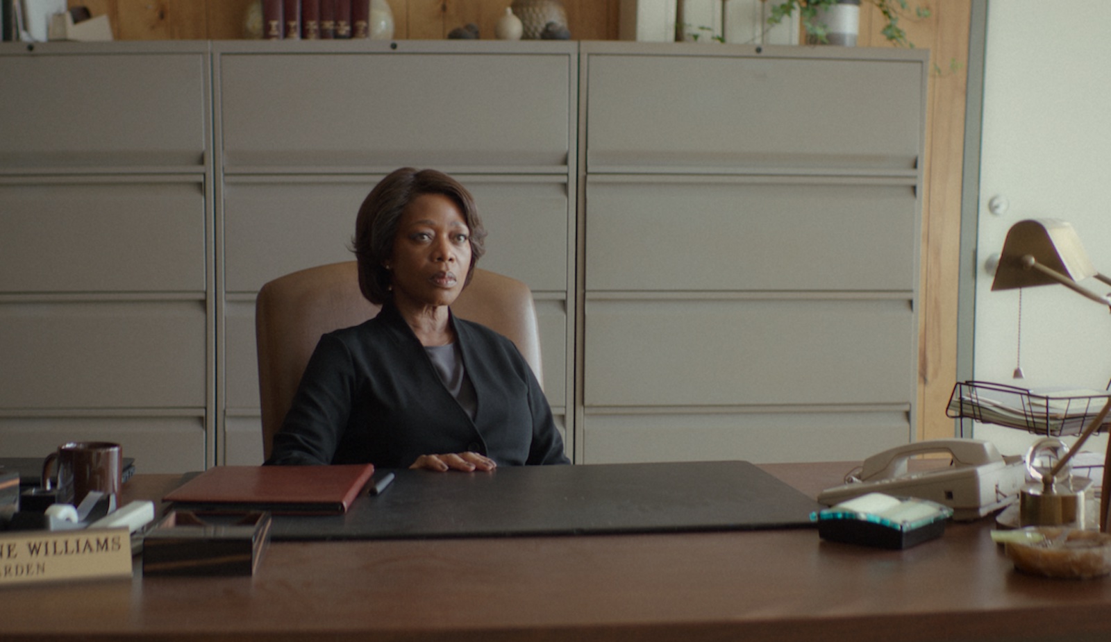 A woman with a serious expression and a black suit sits behind a desk in a drably colored office