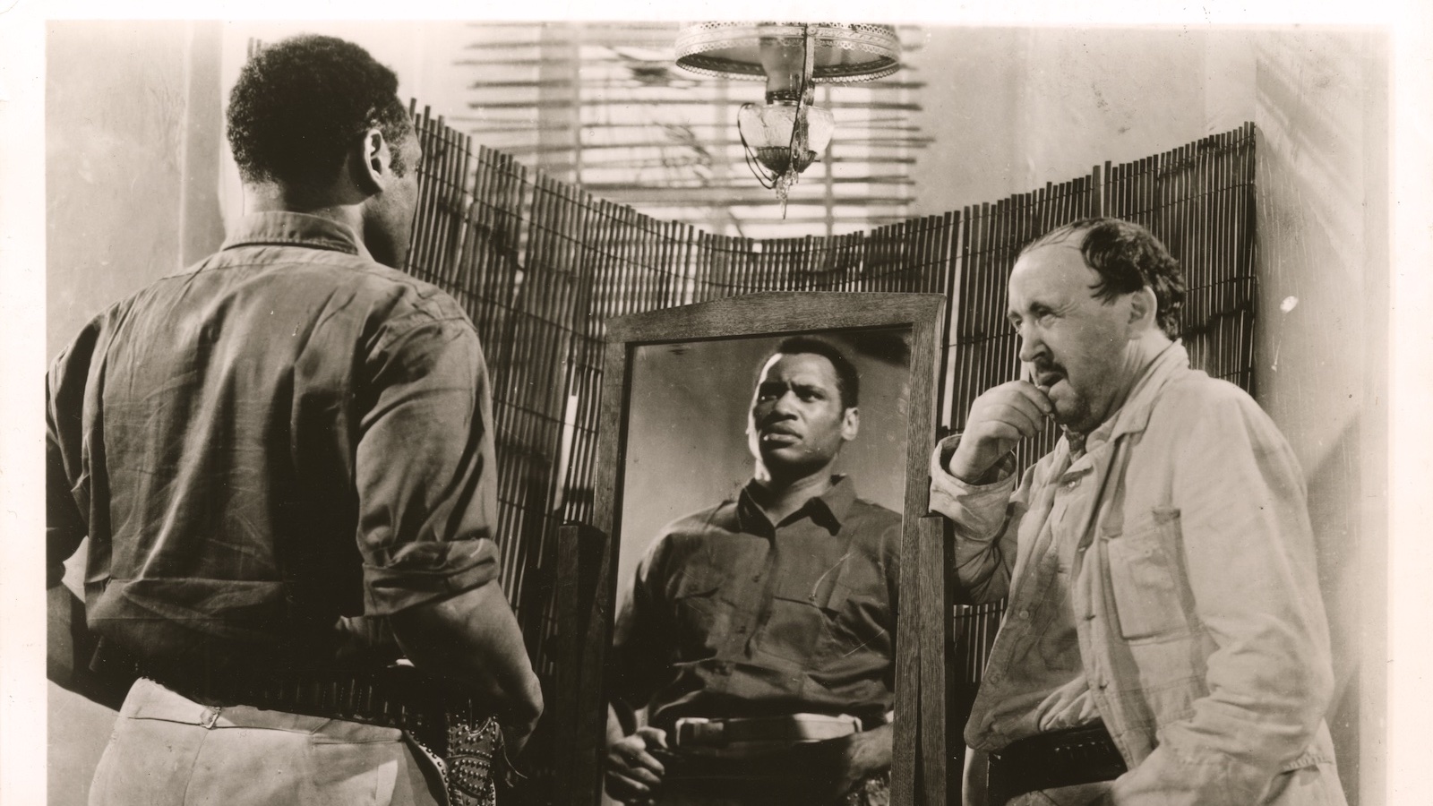 A man looks in a mirror, hands on hips, while another man looks at him from the left
