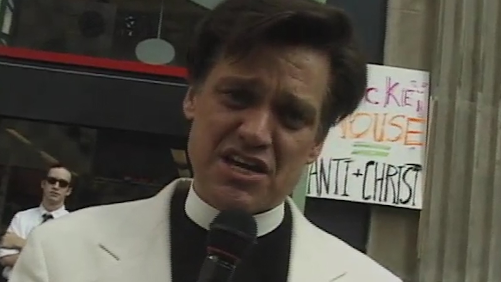 A priest on a sidewalk in the city looks in the camera and talks into a mic