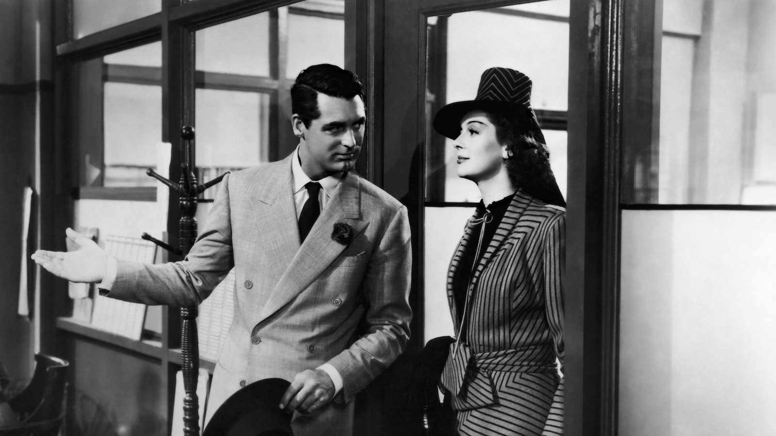 A man in a gray suit welcomes a woman in a pin-striped suit and hat into a newspaper office in a black and white photo