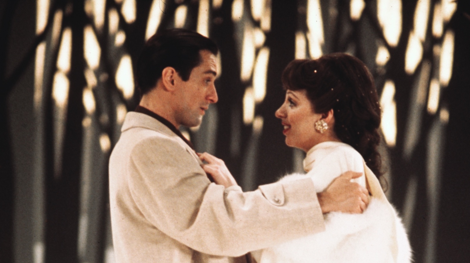 newyork_newyork_5012385.jpg A man in a beige suit and a woman in a white fur coat stand in profile and look at each other, their hands gripping each other, against a forest backdrop