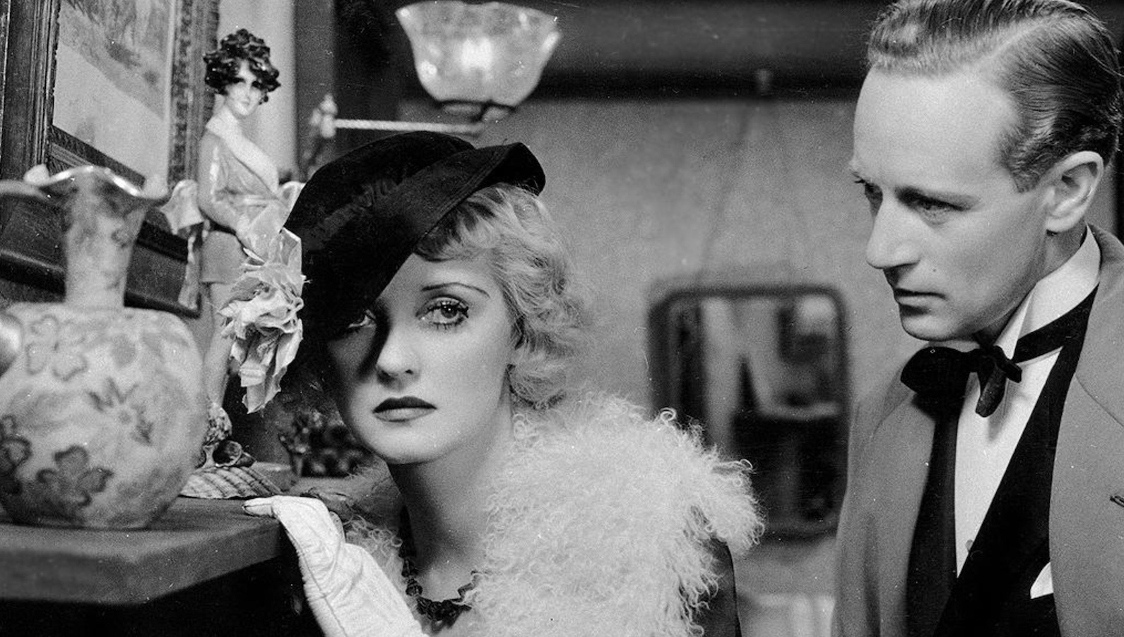 A woman in a hat covering one of her eyes sadly grips the side of a mantel and looks off while a man stands behind her in a black and white image