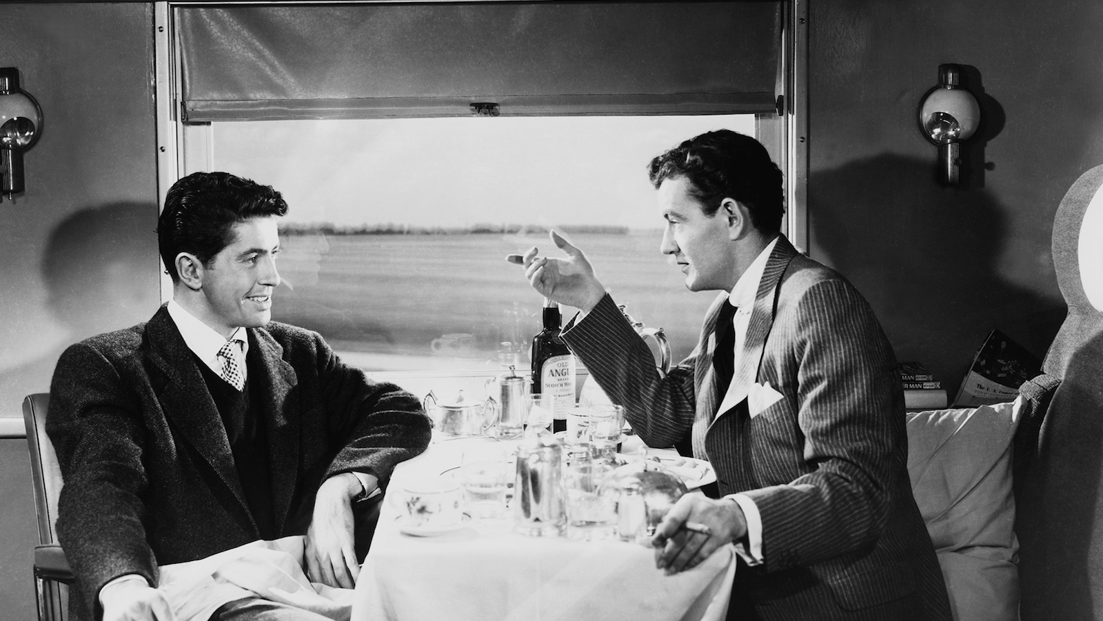 Two men sit at a table on a train compartment, talking pleasantly while landscape goes by in the back of the black and white image