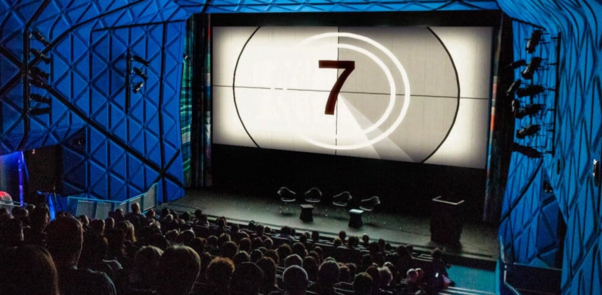 A movie theater with a full audience looking at a screen with the number 7 on it counting down to showtime