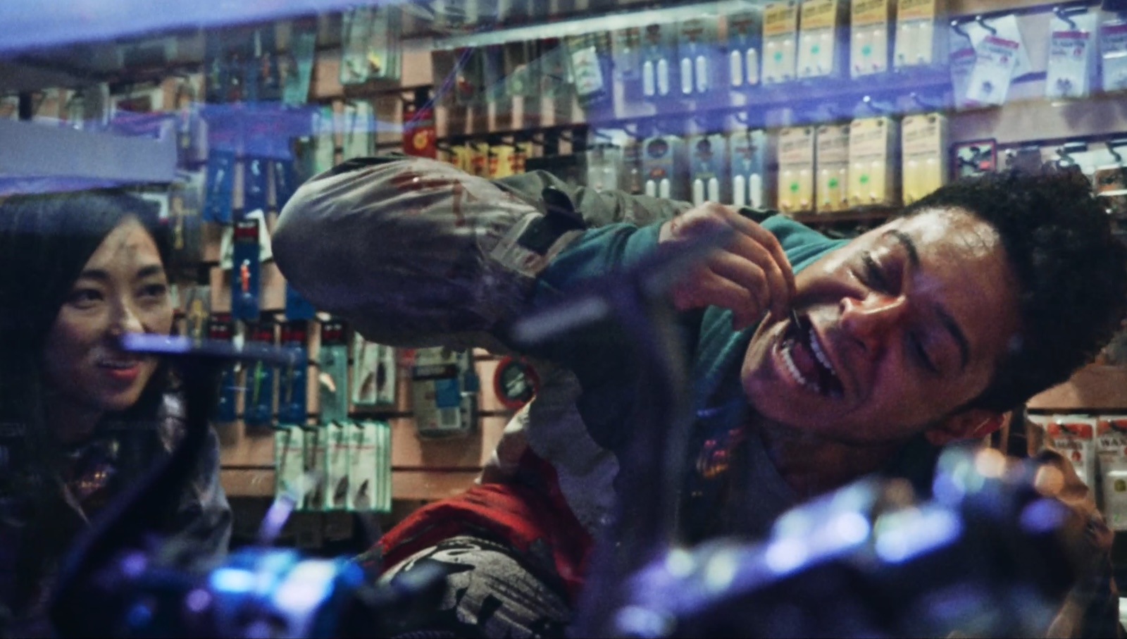 A man tilts his head and manically bites something while a woman watches from the corner against the backdrop of a convenience store
