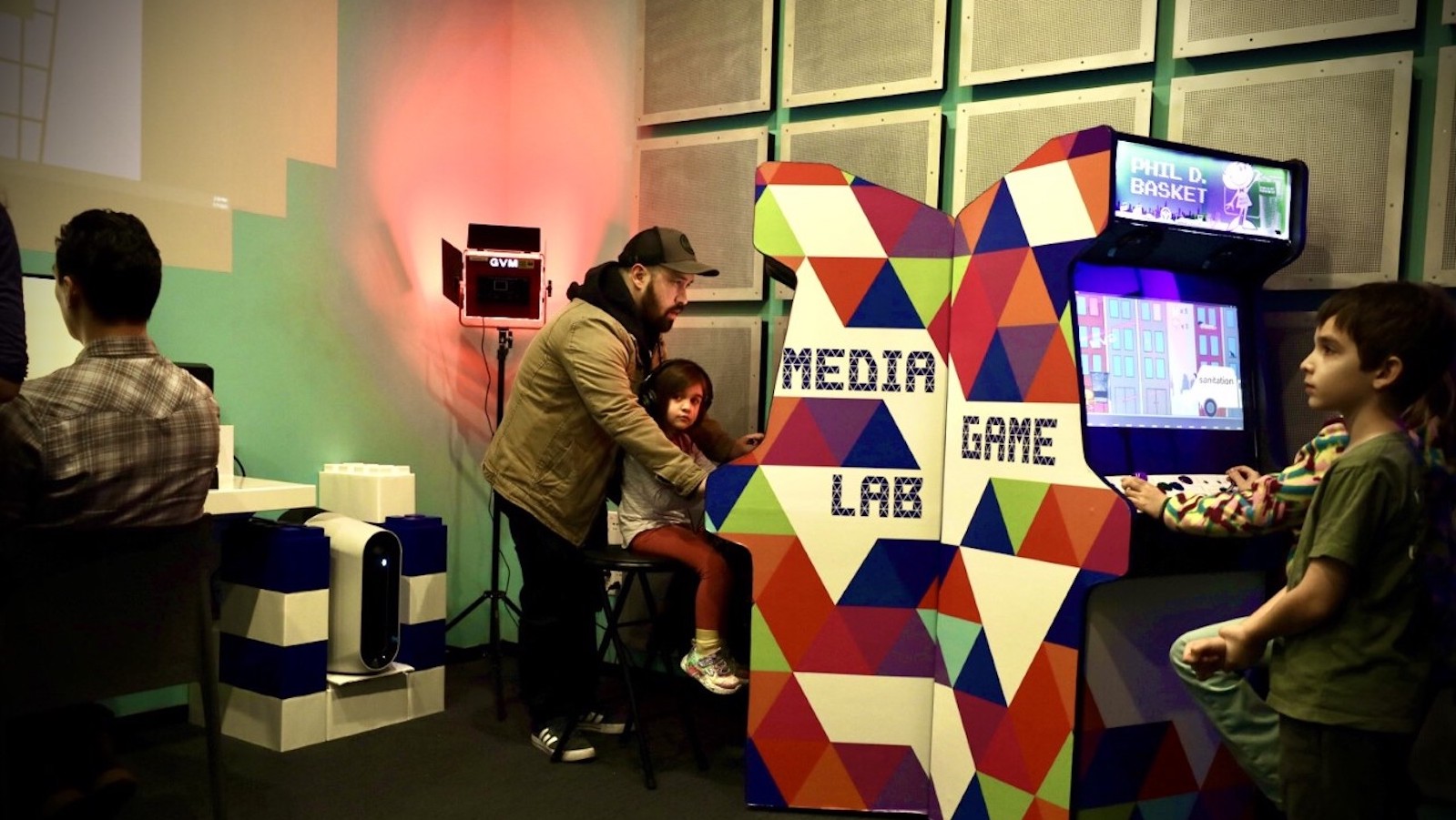 A father and daughter sitting at a video game arcade console; the side of the arcade console says Media Game Lab.