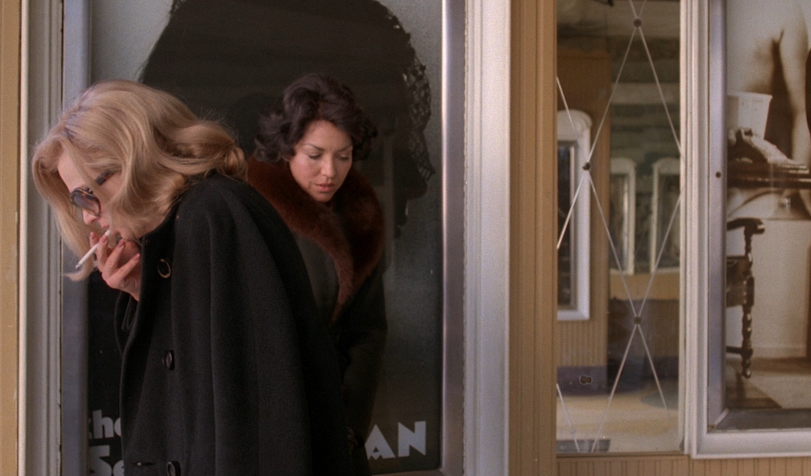 A woman stands in profile with sunglasses on smoking a cigarette, her head down, while another woman stands behind her in the other direction