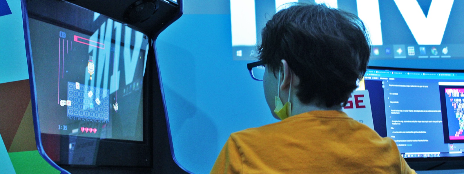 A young person wearing glasses, a yellow shirt, and a facemask dangling from their ears, sits in front of an arcade game, their back to the camera.
