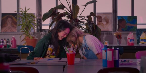 Two friends smile and laugh while sitting in a school room in front of a craft table, their heads on each other's shoulders