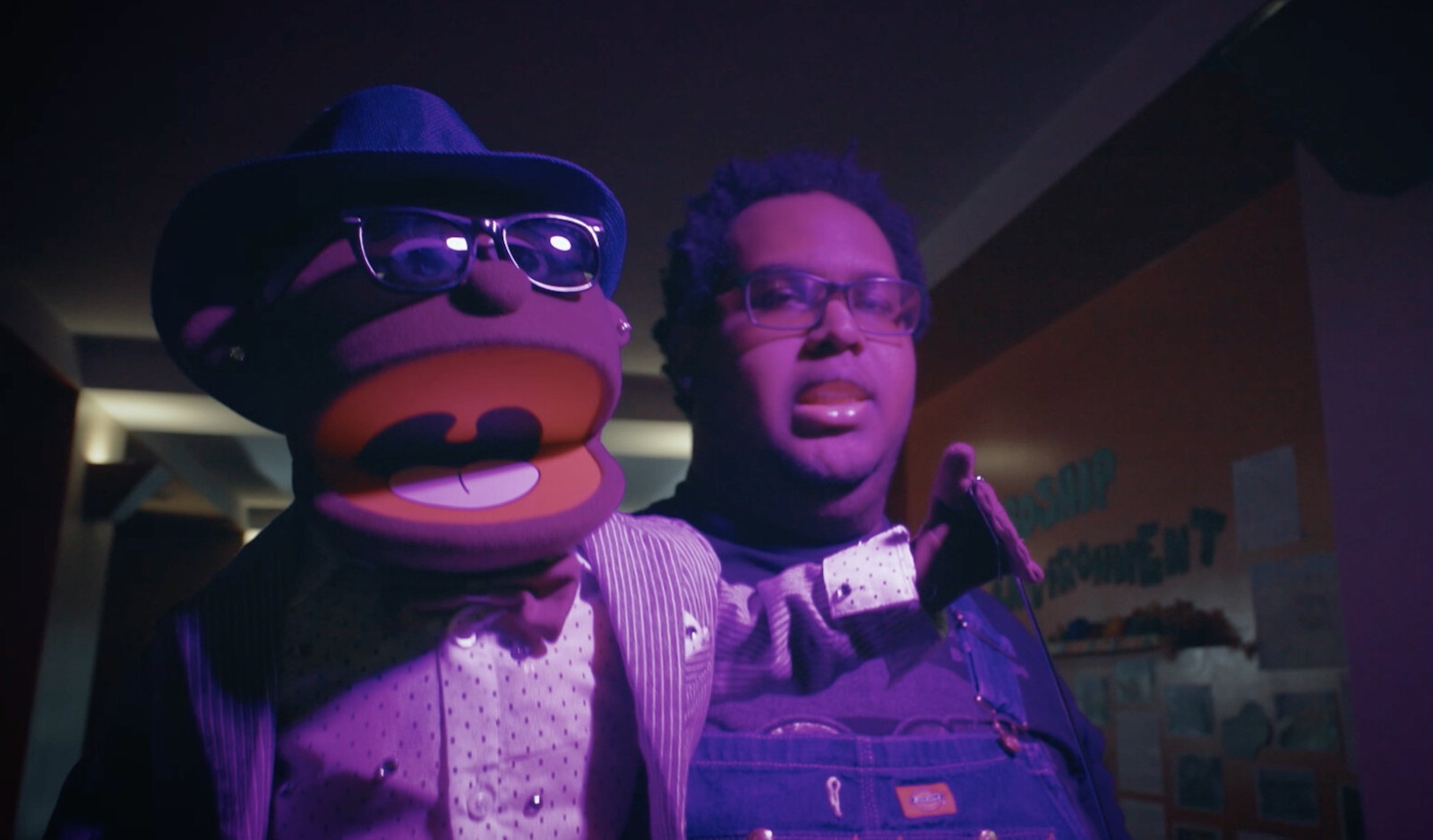A person holds a puppet in the foreground, with a purple light shining on them; the puppet wears a hat and sunglasses and its mouth is open