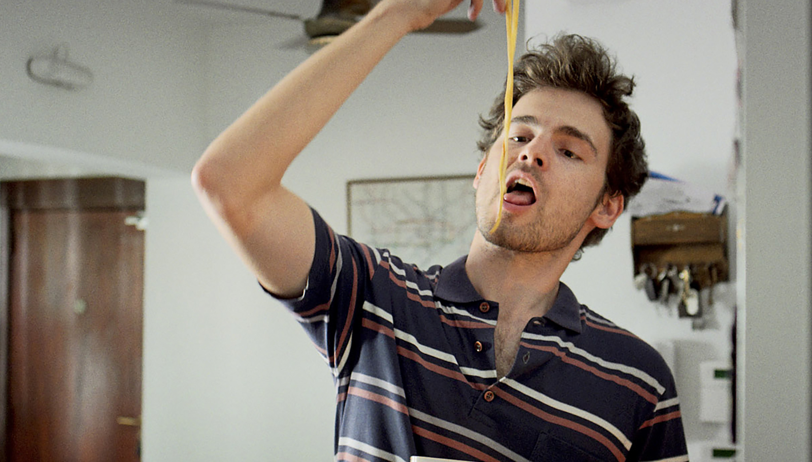 A man in a striped shirt dangles strands of spaghetti into his mouth with his fingers.