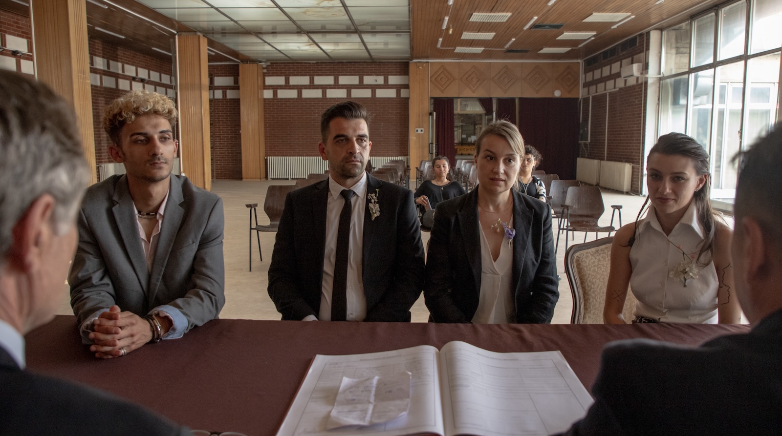 Four people sit before a table in a conference room; the man and woman in the middle are wearing suit jackets and flowers in their lapels as though getting married, and the younger boy on the left and girl on the right appear to be witnessing to their signing legal marriage papers, which are laid out in front of them.