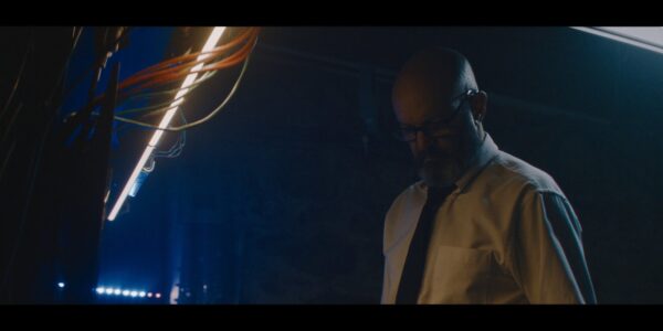 A man in glasses looks down in a dark room with some exposed wiring hanging by a light on the ceiling on the left of the frame