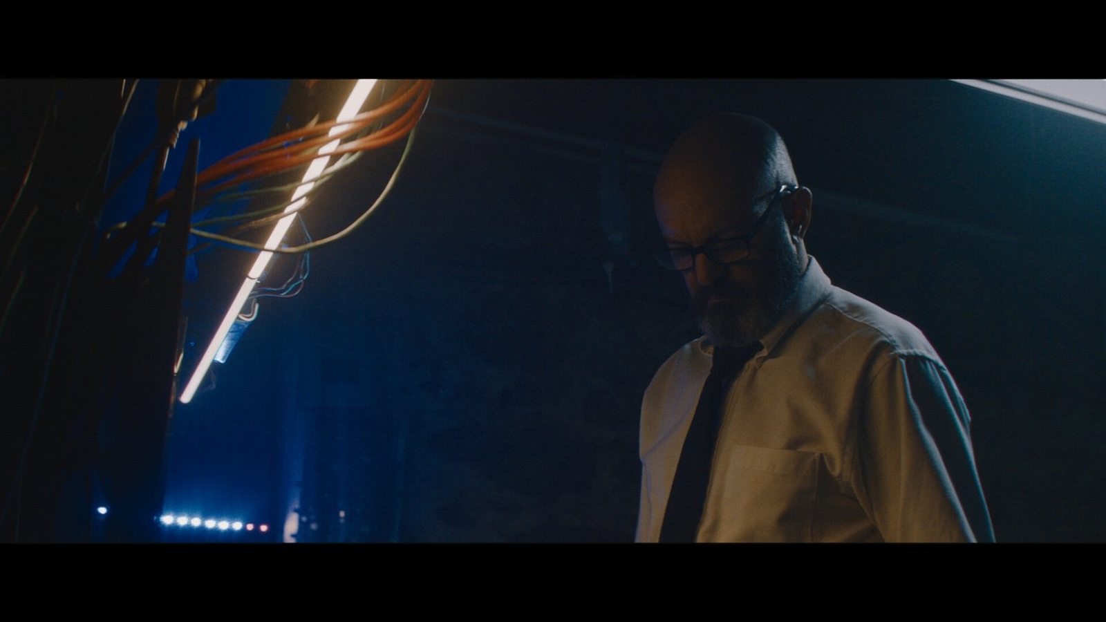 A man in glasses looks down in a dark room with some exposed wiring hanging by a light on the ceiling on the left of the frame