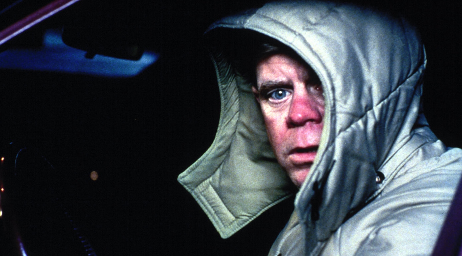 A man in a hooded coat turns to the camera while sitting in a car