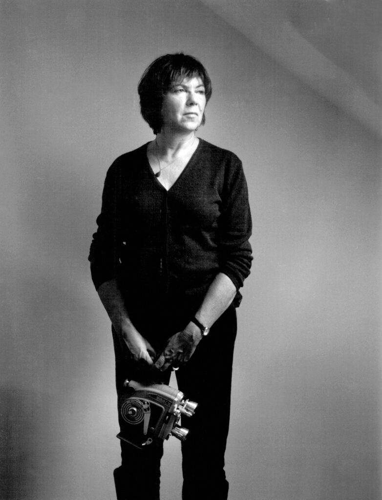 black and white image of a woman holding a camera, dangling from her hands, looking off-screen with a serious expression