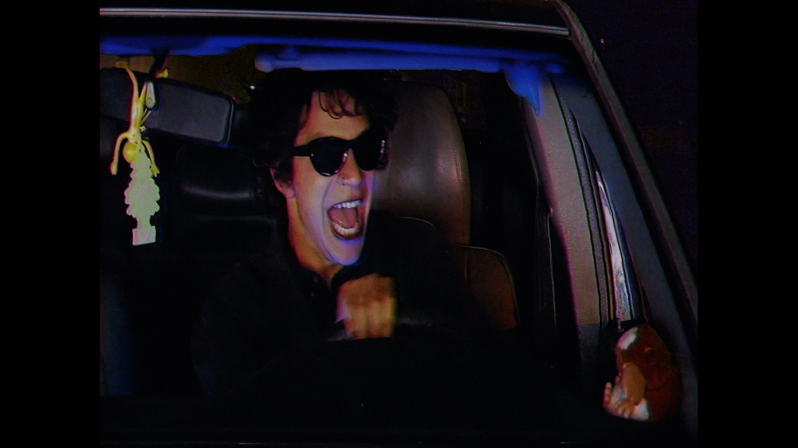 Person in sunglasses screams with mouth open while driving a car at night