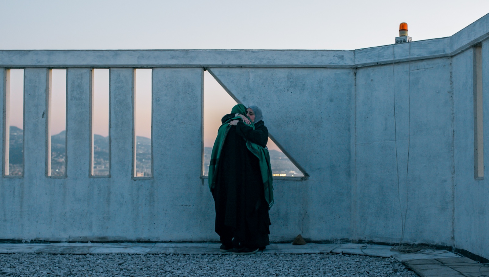 Two women wearing hajibs hug each other while standing on a rooftop, in front of a triangular window, through which we can see a vast mountainous landscape at dusk.