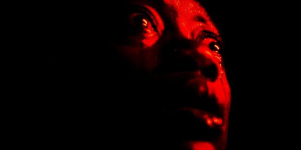 An intense close-up of a face drenched in red looking up and off-screen in panic