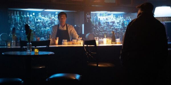 A bartender in an apron wears a fearful grimace as he greets a disheveled patron, whom we only see from behind as he approaches the bar