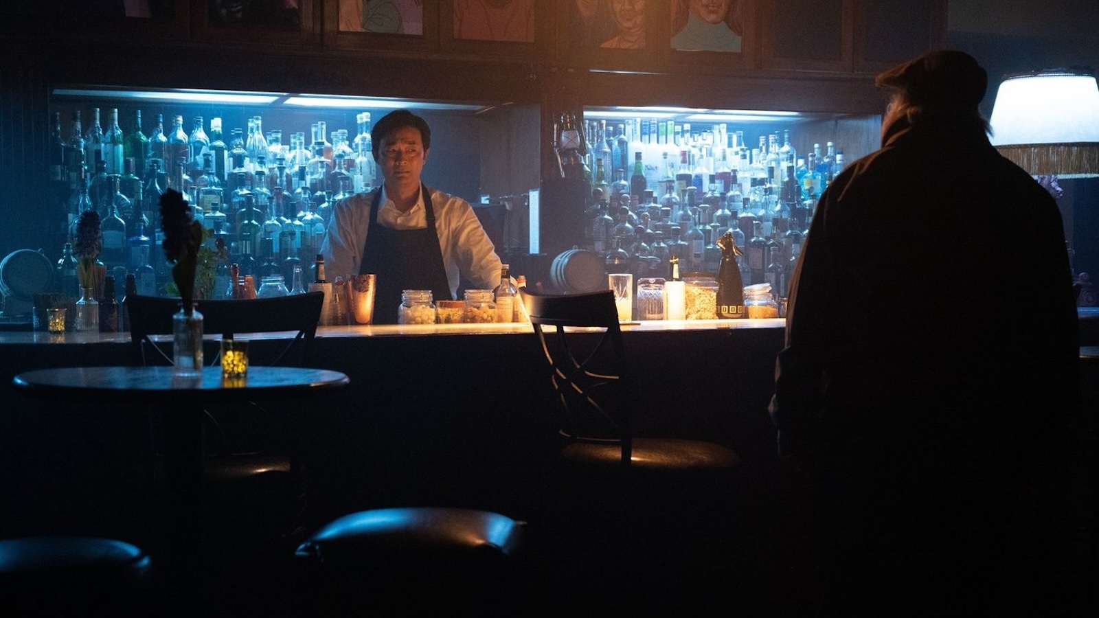 A bartender in an apron wears a fearful grimace as he greets a disheveled patron, whom we only see from behind as he approaches the bar