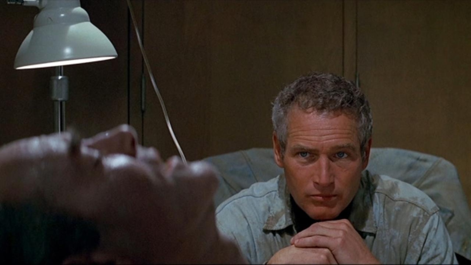 A man with gray hair sits in a hospital, his hands under his chin pensively, as he stares at a man with his eyes closed and mouth open in the foreground.