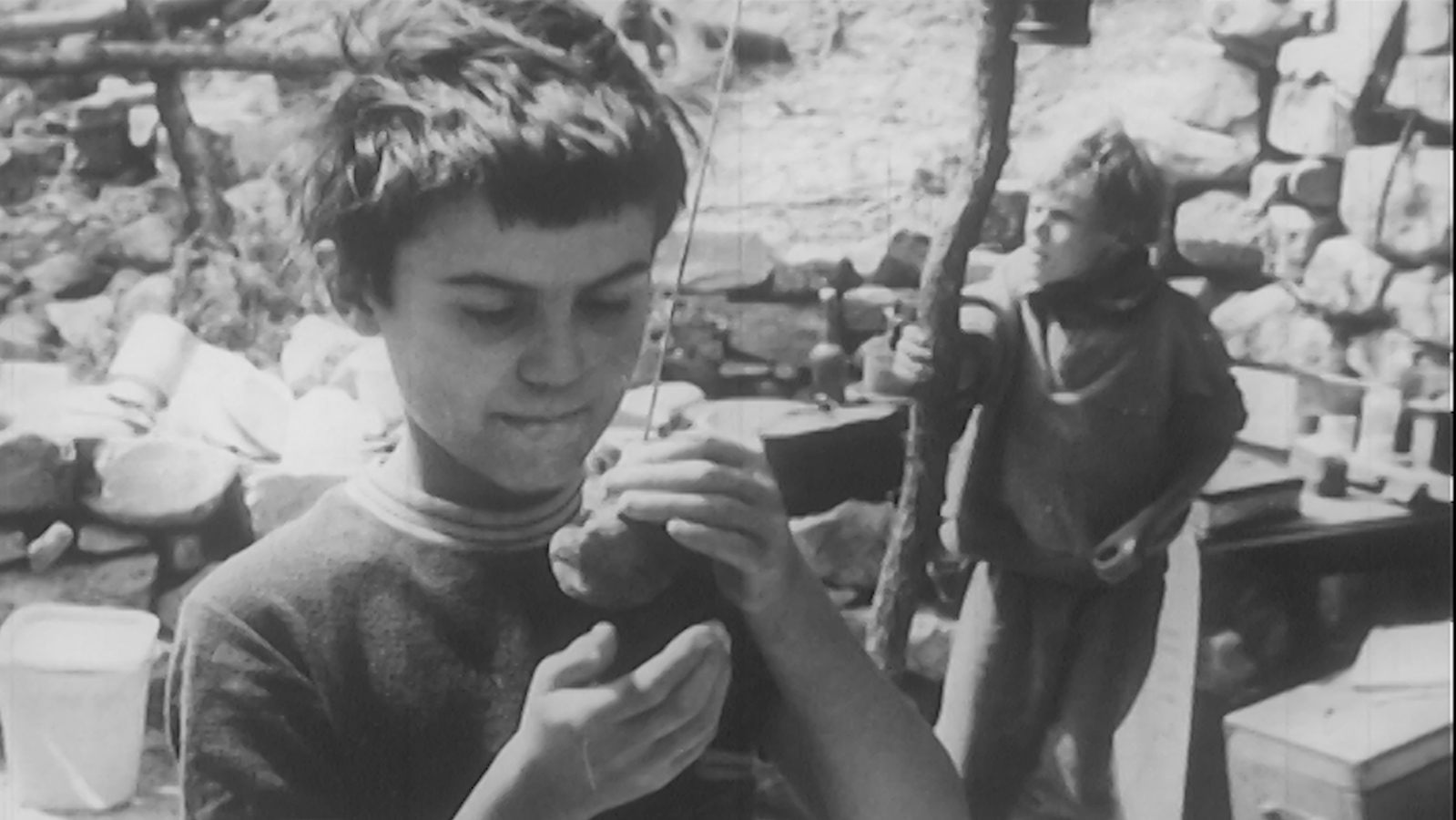 Ce-gamin-la_08 a black and white image of a young boy standing outside cupping an object in his hands, while another boy behind him clutches a wooden post