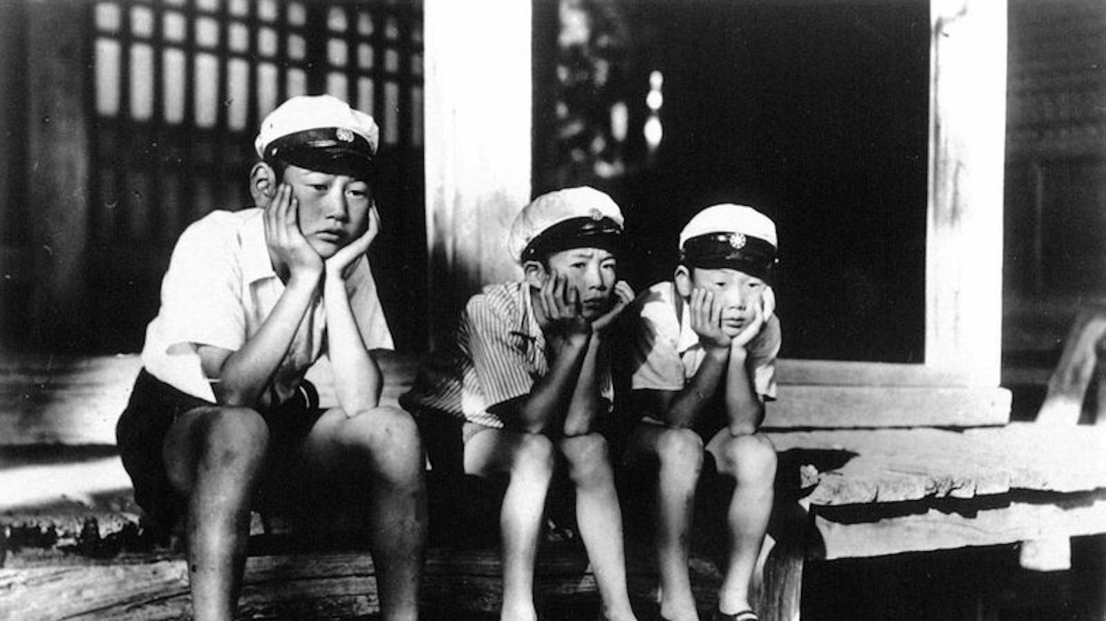 A black and white photo of three Japanese boys in white caps sitting forlornly with their chins in their hands on the edge of a house's front porch.