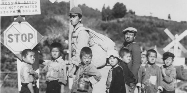 A black and white image of a group of Japanese children and two adults standing in front of a STOP sign at the side of the road looking at the camera forlornly