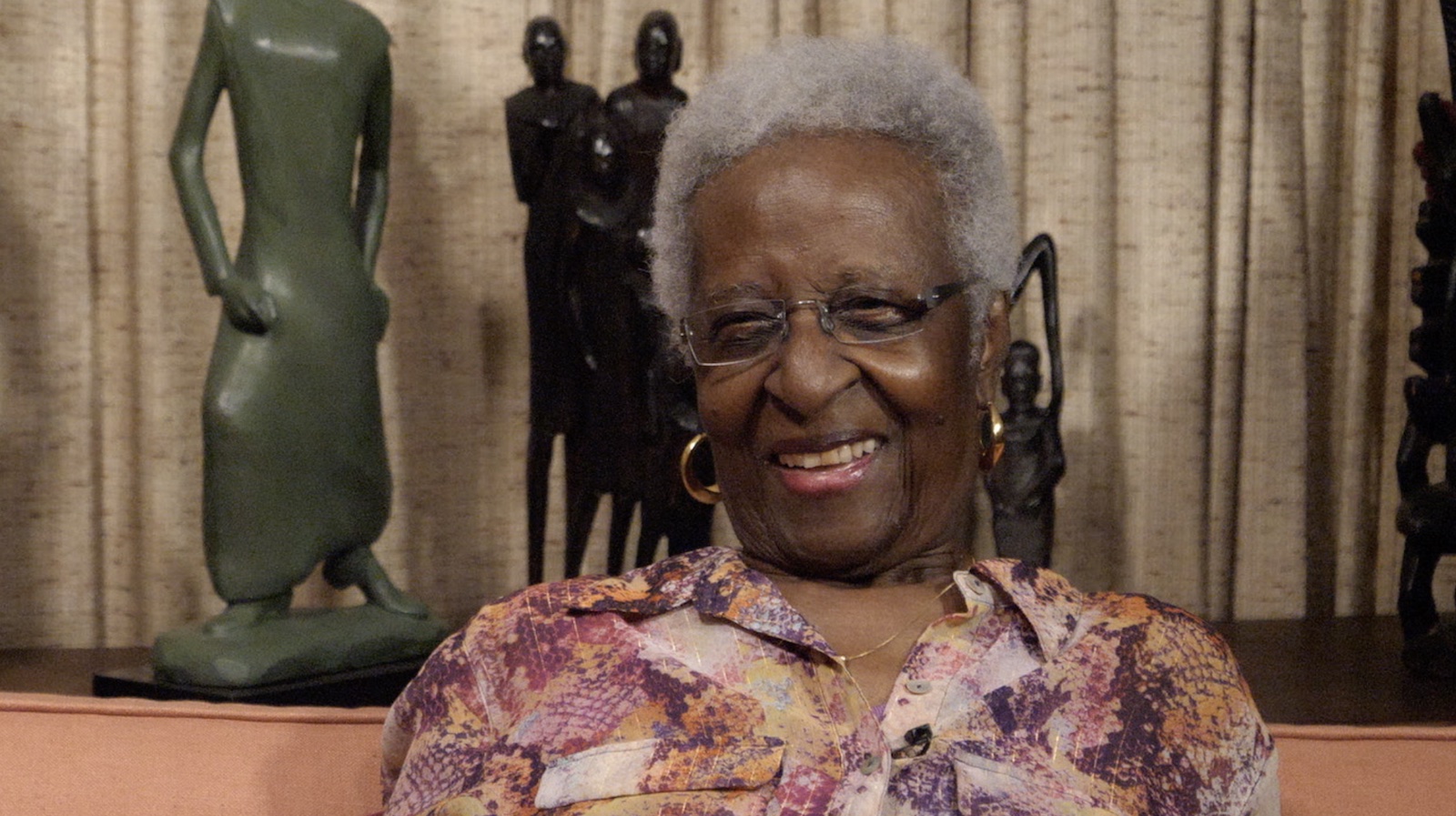 An elderly woman with dark brown skin, white curly hair, gold hoop earrings, and a purple and orange colored shirt sits and smiles, talking to camera, in front of green and black African-style sculptures