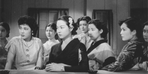 A black and white image of a group of six Japanese women looking left off-screen, the womanin front leaning her elbow on a table, all look serious