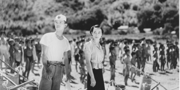 A black and white photo of a Japanese man and woman standing on the shore of a beach, looking off, as lines of children in formation in the background of the shot face the water
