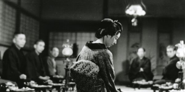 A black and white photo of a woman in traditional Japanese clothes, seen from the back, leaning over a table setting on the floor, which human figures out of focus in the background.