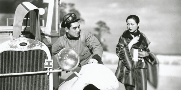 A black and white photo of a man in a cap polishes the side of the front wheel of a bus while a woman closely clutching her coat looks on.