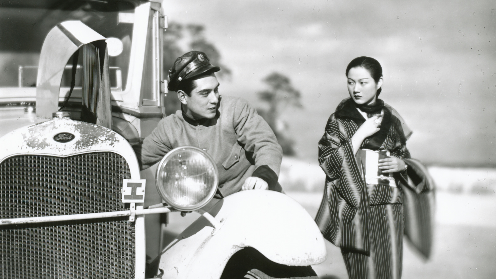 A black and white photo of a man in a cap polishes the side of the front wheel of a bus while a woman closely clutching her coat looks on.
