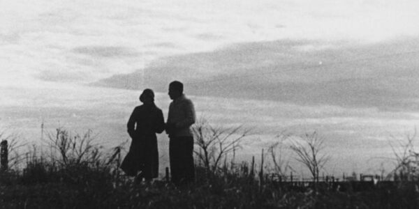 A black and white image of two human figures silhouetted against the sky standing amidst tall grasses