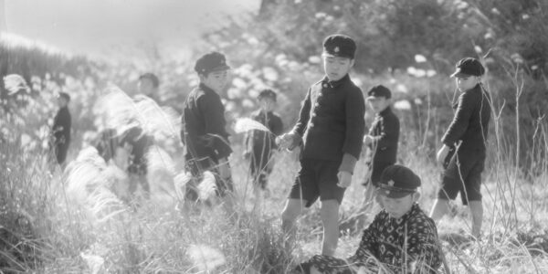 A black-and-white image from the 1930s of young Japanese boys dressed in school uniforms and caps standing amidst a sun-dappled field of wheat