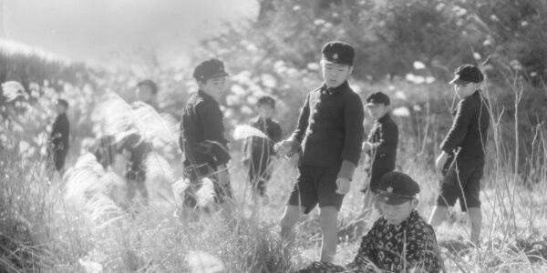 A black-and-white image from the 1930s of young Japanese boys dressed in school uniforms and caps standing amidst a sun-dappled field of wheat