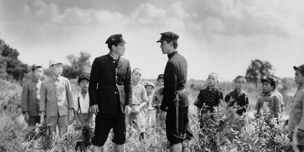 A black and white photo of two men in hats and uniforms standing and facing each other in a field of grass amidst other children figures who surround and watch them.