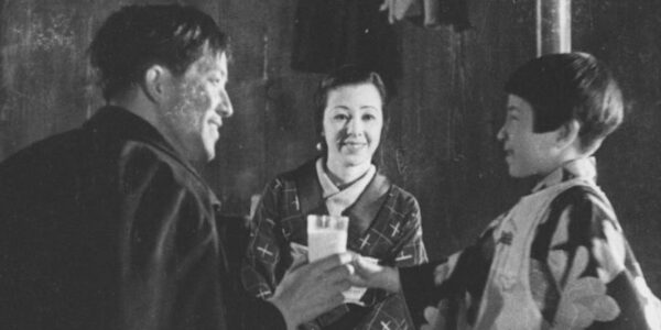 A woman stands between a man and woman as they look at each other and exchange a glass of milk