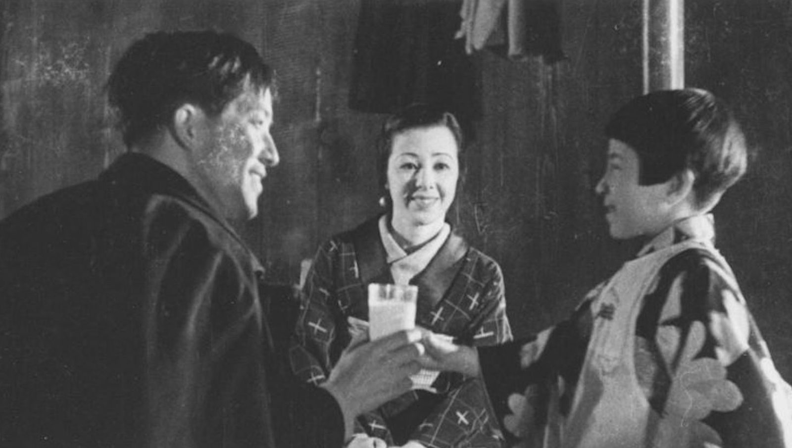 A woman stands between a man and woman as they look at each other and exchange a glass of milk