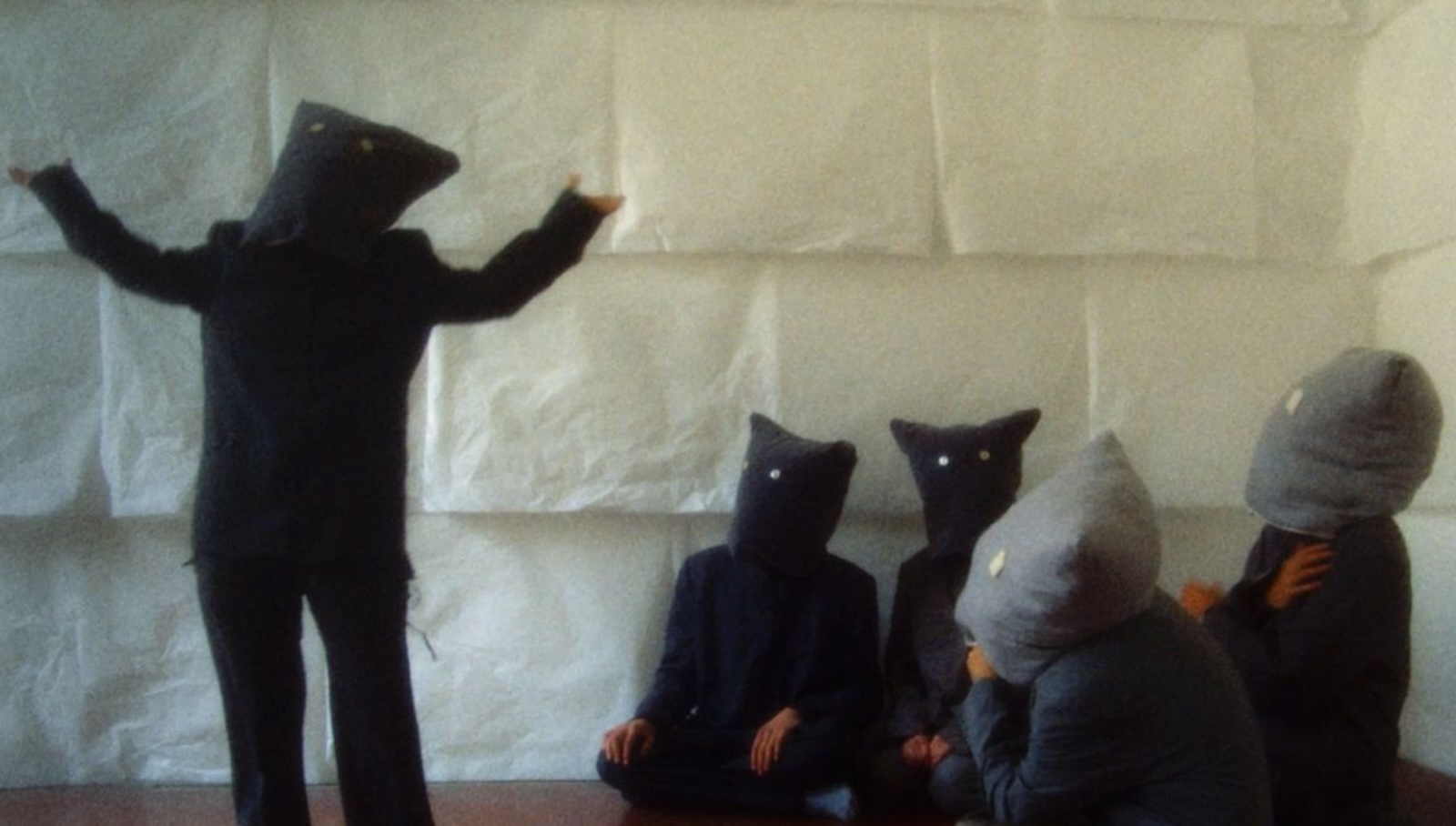 A group of five people with bags over their heads and buttons for eyes in a room with white walls. One of the people stands with their arms outstretched and the others are sitting on the floor