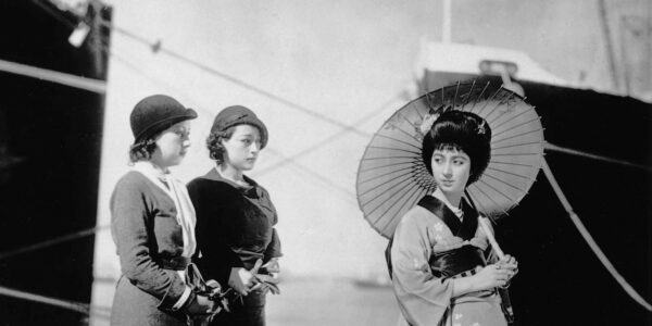 At a seaside dock, two Japanese women in modern clothes and hats look at a woman in traditional geisha clothes and parasol