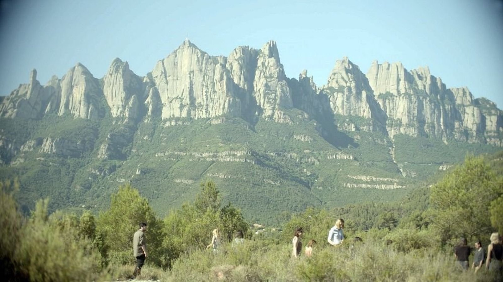 A vast mountainous landscape, with people in the foreground standing amidst green trees and bushes.