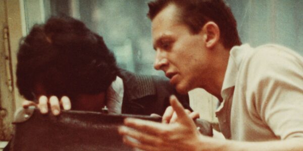 A man gestures aggressively at a woman, who hides her head behind her bag as they sit in public.